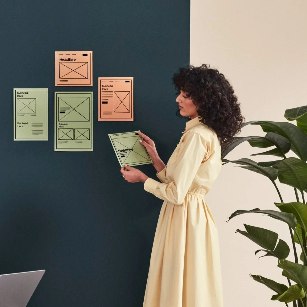 A woman in a light yellow dress appears to be a graphic designer or marketer, examining and arranging color-coded and wireframe layout papers on a dark blue wall, with a laptop visible in the lower corner and large green leafy plant on the right side.