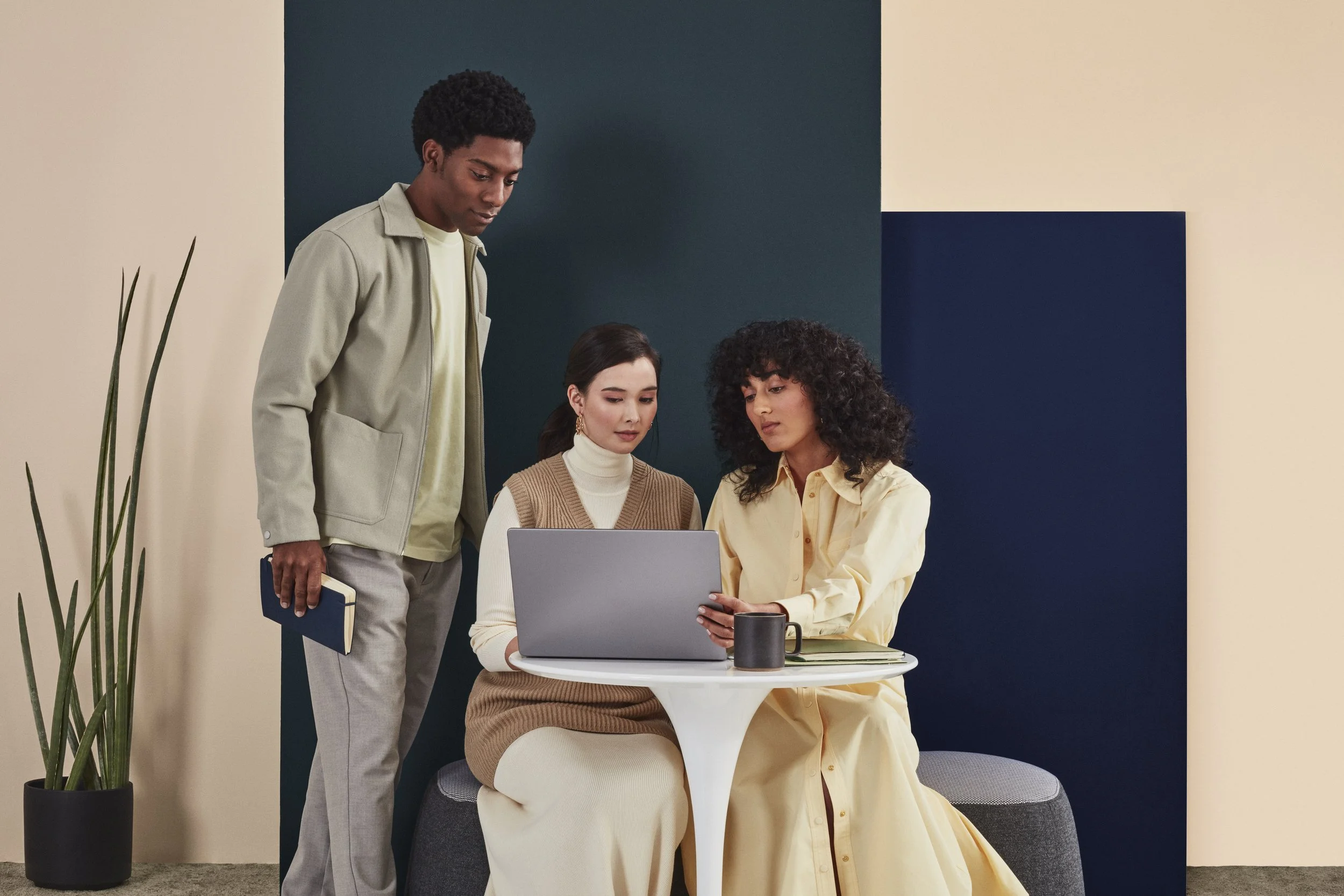 Three young women collaborating around a laptop in a modern office space with a dark green wall background, a coffee mug, and a notebook on the table.