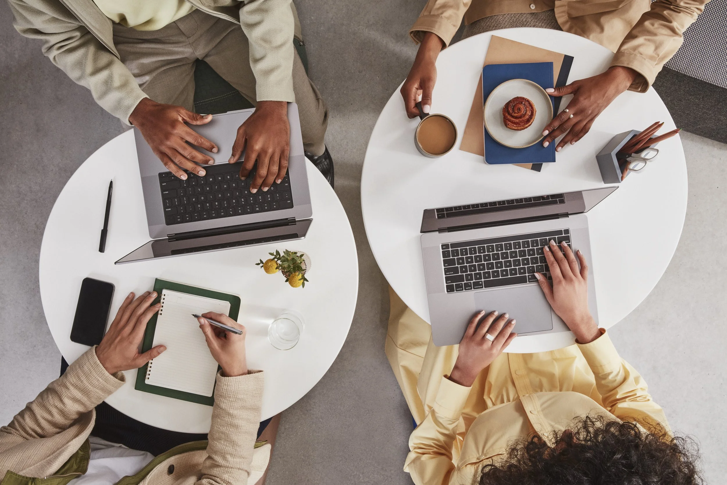 Top-down view of three people working at two white round tables with laptops, notebooks, pens, coffee, and snacks.