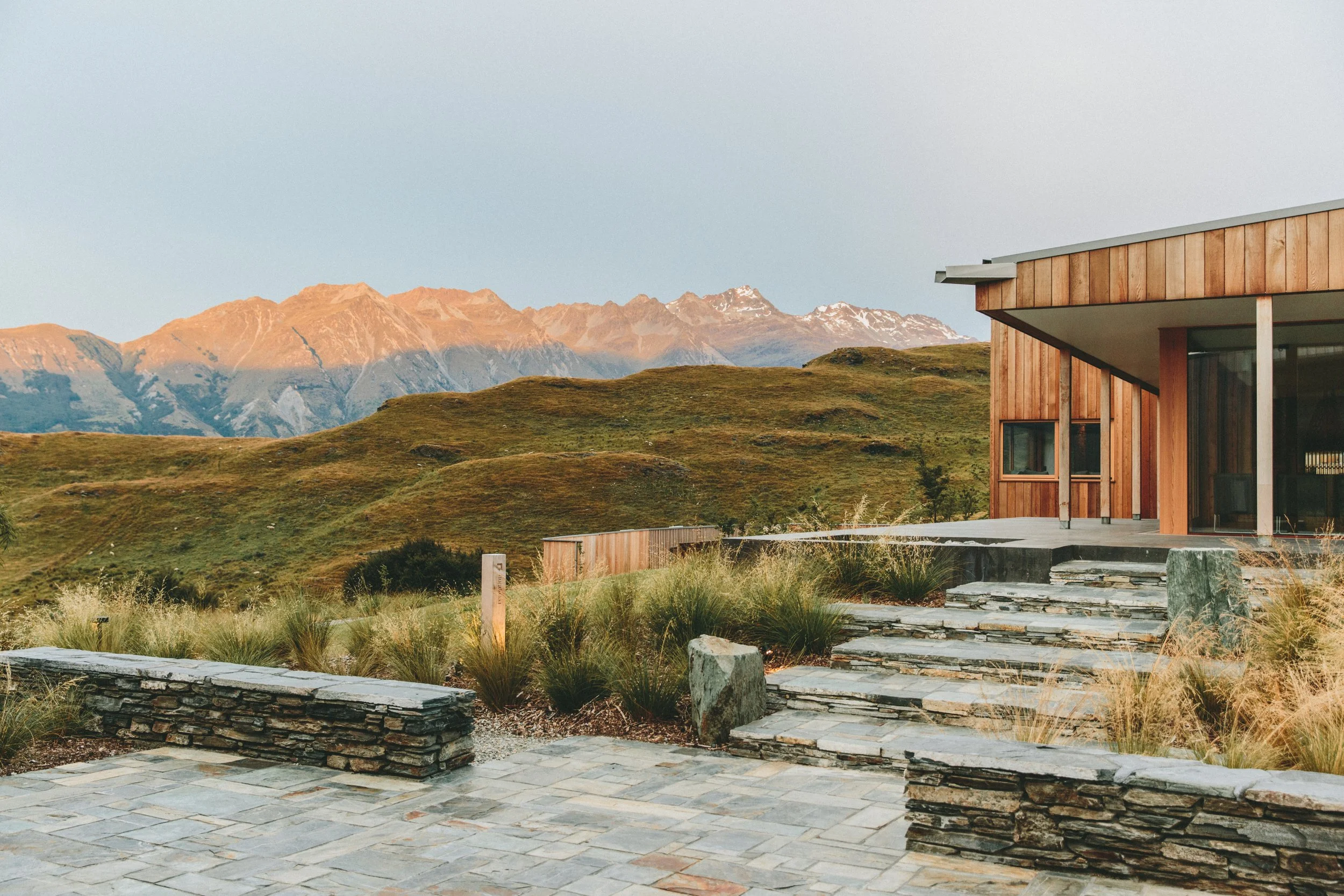 Modern house with wooden exterior on a landscaped stone patio, overlooking green hills and mountains at sunset.